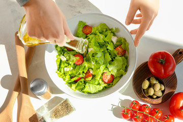 Woman adding olive oil into bowl with fresh vegetable salad on table