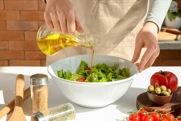 Woman adding olive oil into bowl with fresh vegetable salad on table