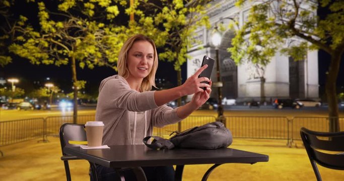 Caucasian woman sitting outside at cafe in Paris taking selfie to send to friend, Young blonde female taking photo of herself near the Arc de Triomphe on smartphone, 4k