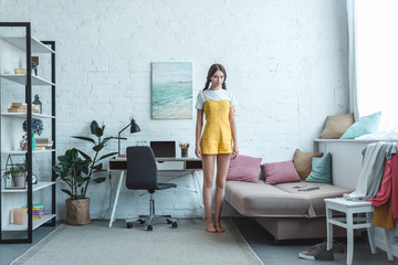 Fototapeta premium beautiful teen girl standing in living room with sofa, bookshelf and laptop on table