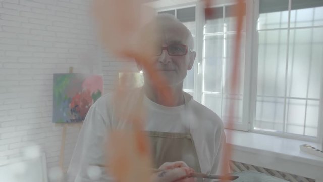 Artist painting in his atelier using orange paint and brush. Wearing white t-shirt and beige apron, eyeglasses. Big white windows and other colorful art paintings behind him. Looking inspired .
