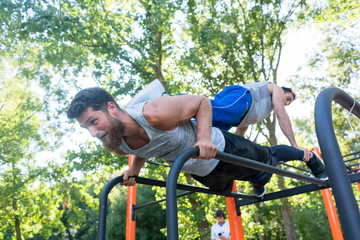 Low-angle view of a strong young man supporting his friend on his back, while doing extreme pull-ups during partner workout in a calisthenics park in summer