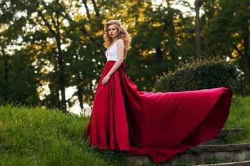 Fototapeta premium Fashion style photo of young beautiful woman in evening dress with white top and red flying skirt posing in summer park at sunset under green trees and close to old brick tower.