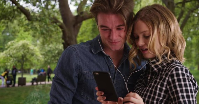 Young millennial couple at the park watching funny baby videos on smartphone, Caucasian male and female sharing headphones, relaxing outdoors, 4k