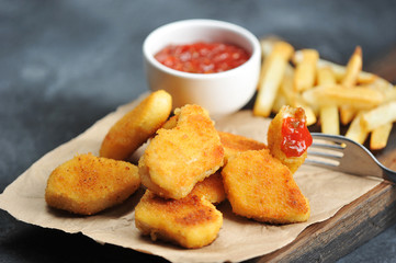Chicken nuggets and French fries on a wooden board. Next cup with ketchup. One of the nuggets is pinned on the fork. Close-up. Gray background. Macro photography.