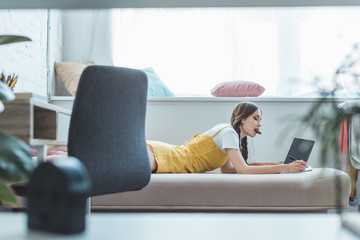 teen girl eating cookie writing in copybook and using laptop on sofa near window