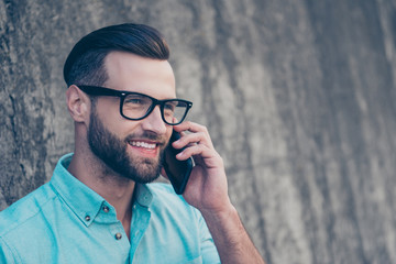 Portrait with copyspace of cheerful glad man with modern hairdo in blue shirt speaking by smart phone with friend having beaming smile isolated on grey stone wall. Advertisement concept