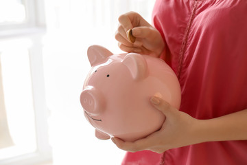 Woman putting coin into piggy bank indoors. Money savings concept