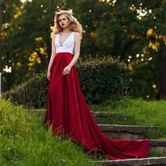 Obraz na płótnie Canvas Fashion style photo of young beautiful woman in evening dress with white top and red flying skirt posing in summer park at sunset under green trees and close to old brick tower.
