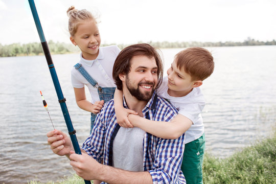 A picture of happy family standing together at river shore. Boy is hugging dad while man is looking at him and smiling. Also guy is holding fish-rod. Girl is standing behind father and looking at him.