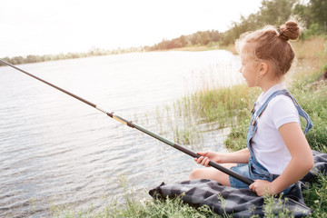A picture of girl sitting alone at river shore. She is fishing. Girl is holding fish-rod with both hands. She is looking at water. Girl is very serious.