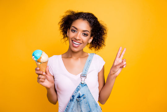 Portrait of cheerful positive girl gesturing v-sign having vanilla ice cream in waffle cone with caramel isolated on yellow background. Flavor dairy healthy nutrition concept