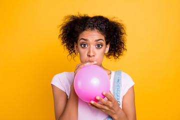 Portrait of amazed astonished girl with wide open eyes blowing up pink balloon preparing surprise celebrating birthday isolated on yellow background