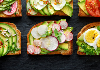 Avocado sandwiches, toasts with various vegetarian toppings on a black stone background, top view