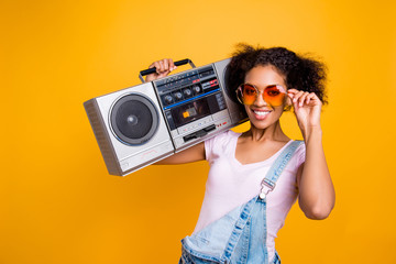 Portrait of fancy toothy girl with beaming smile in eyewear holding boom box on shoulder looking at camera isolated on yellow background. Music lover fan hobby concept