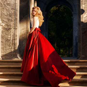 Fashion style photo of young beautiful woman in evening dress with white top and red flying skirt posing at stairs at sunset