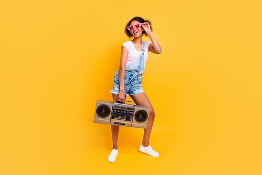 Full size portrait of cheerful positive girl in eyewear jeans overall holding boom box in hand going to make party isolated on yellow background. Music lover fan concept