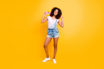 Fototapeta na wymiar Fullbody portrait of cheerful pretty chick in jeans overall holding skate board on shoulders isolated on yellow background. Extreme balance concept