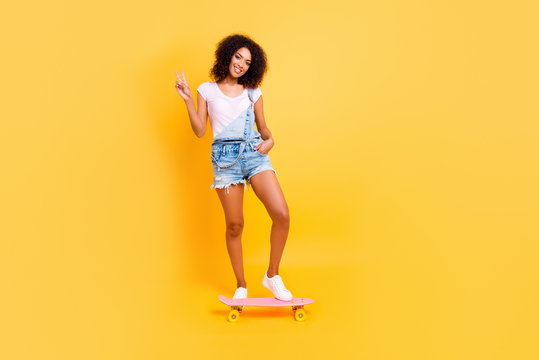 Full body portrait of peaceful friendly girl in denim overall riding on skate board gesturing v-sign holding hand in pocket isolated on yellow background. Activity athletic extreme balance concept