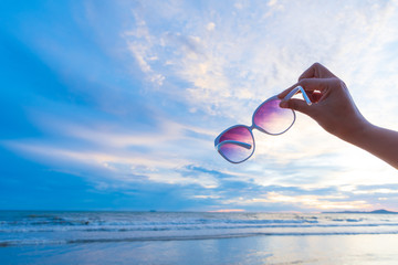 Soft focus on woman hand holding sunglasses over sea and Sandy beach in background during sunset...