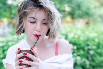 Attractive girl with red lips and manicure is sitting in the park and drinking coffee