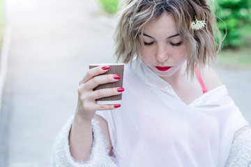 Attractive girl with red lips and manicure is sitting in the park and drinking coffee