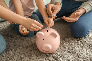Little girl with her parents sitting on carpet and putting coin into piggy bank indoors. Money...