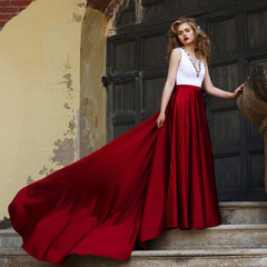 Young beautiful woman standing at stairway and posing in evening dress with white top and red...