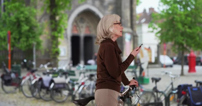 Happy older Caucasian woman on bike using smartphone while on vacation in Bruges, Smiling senior tourist in city texting on mobile phone, 4k