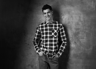 Portrait of young trendy handsome man with short dark hair wearing checkered shirt and brown trousers standing and posing against gray concrete wall