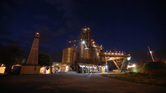 Night panorama of loading grain crops on bulk freighter ship via trunk to open cargo holds at silo terminal in seaport from trucks. Cereals bulk transshipment. Transportation agricultural products.