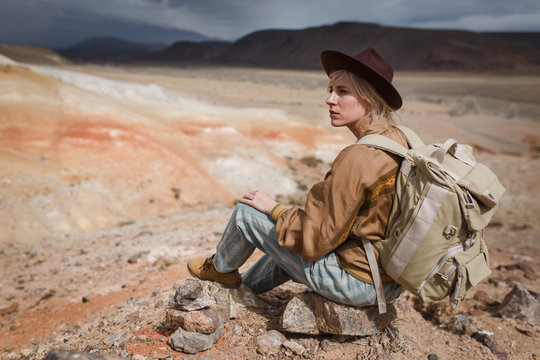 Beautiful young girl in hare, hat and with backpack siting among sands in wild