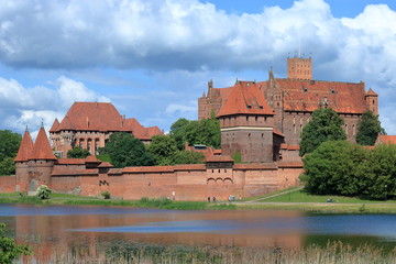 Zamek krzyżacki w Malborku, Polska, rzeka Nogat, zieleń wokół murów budowli, malownicze chmury na niebieskim niebie - obrazy, fototapety, plakaty