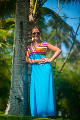 Beautiful fashionable young woman in dress and sunglasses standing near palm trees and smiling