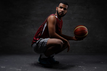 African-American basketball player in sportswear squatting with a ball on a dark background. 