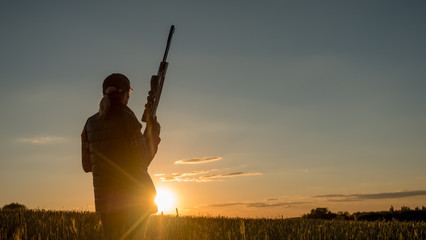 Silhouette of woman hunter. It stands in a picturesque place with a gun at sunset. Sports shooting and hunting concept - obrazy, fototapety, plakaty