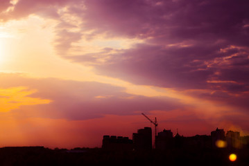 Fototapeta premium Magenta sunrise above silhouette of city buildings with tower cranes. Unusual cityscape with beautiful dawn. Picturesque warm city landscape with bright sky. Natural glare from sun (lens effect).