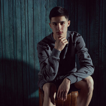 Thoughtful young handsome man with dark short hair sitting against green wooden wall in dark like and looking at you or in camera