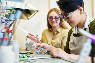 Side view portrait of two modern female artists working with glass, focus on ypung Asian woman in glassworking studio