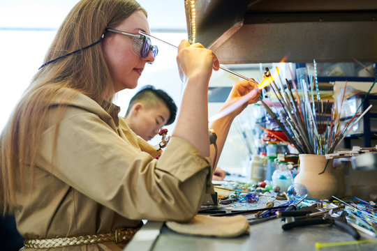 Side view portrait of young woman melting glass over gas torch while making handmade beads in modern creative workshop, copy space