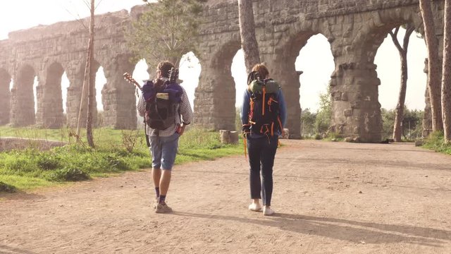 Young couple backpackers tourists walking near roman aqueduct arches in parco degli acquedotti park ruins in rome at sunrise with guitar and sleeping bag slow motion steadycam