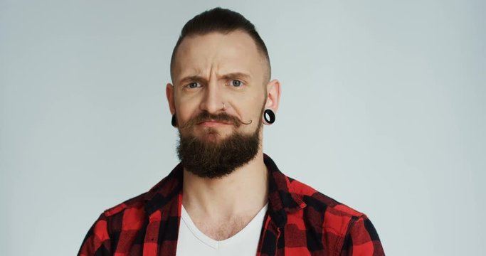 Portrait of the Caucasian hipster man in the plaid shirt and with earings smiling and then doing funny strange face on the white wall background. Close up. Indoors.