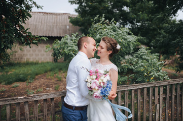 Beautiful and smiling newlyweds are hugging in the village, against the backdrop of an old house and a wooden fence, in green foliage. A wedding portrait of a stylish bride and a cute bride in nature.