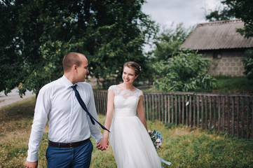 A beautiful bride and a stylish groom are walking, holding hands on the background of an old house and a wooden fence, in green foliage. A wedding portrait of a honeymoon couple walking outdoors.