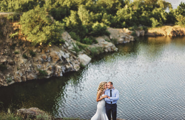A stylish bridegroom embraces a beautiful bride with curly hair from behind, against the background of rocks and a lake. Lovely newlyweds are standing on the edge of the cliff.