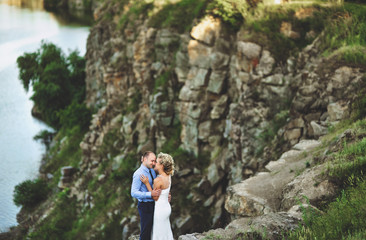 Naklejka na ściany i meble A nice bridegroom embraces a beautiful bride with curly hair and rests her forehead, standing on a precipice. Lovely and smiling newlyweds stand on a rock, against the background of rocks and the sea.