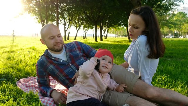 Little baby girl talking on the phone, holding a smartphone in her hands. Happy family having a picnic outdoors in summer at sunset