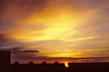 Fototapeta premium Beautiful cloudy dramatic morning sky above silhouette of city buildings. Picturesque dawn in city. Aircraft trail on vivid clouds. Background from unimaginable fantasy sunrise in heaven.