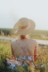 A young beautiful girl in a straw hat sitting back to the camera on the field