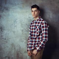 Obraz na płótnie Canvas Portrait of young trendy handsome man with short dark hair wearing checkered shirt and brown trousers standing and posing against gray concrete wall